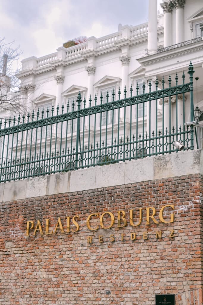Heiraten im Palais Coburg Goldene Lettern „PALAIS COBURG RESIDENZ“ auf einer roten Backsteinmauer, darüber ein grünes Ziergitter und im Hintergrund die weiße Säulenfassade des Palais Coburg.