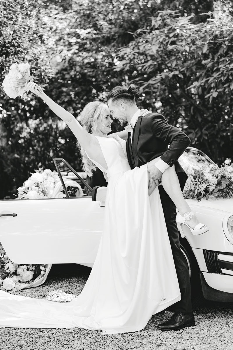 Brautpaar posiert während der Hochzeitsplanung vor weißem Oldtimer im Garten, in einer verspielten und eleganten Pose
