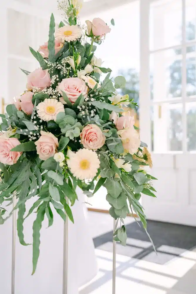 Großes Blumengesteck mit rosafarbenen Rosen, Dahlien und Eukalyptus auf weißem Ständer vor einem Fenster der Orangerie Eisenstadt