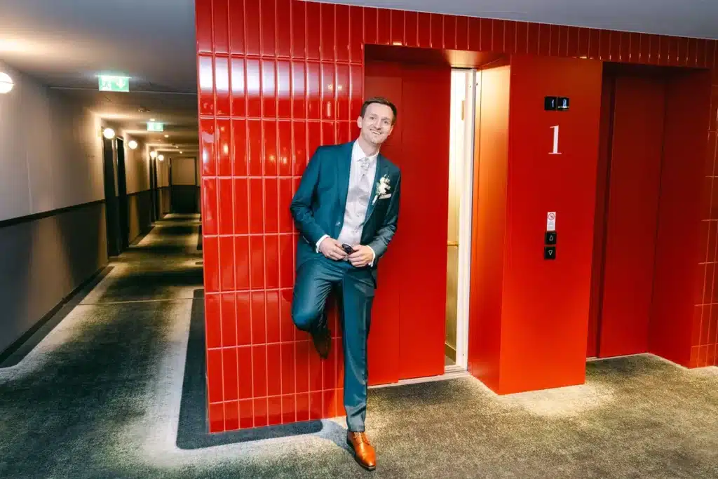Bräutigam im blauen Anzug lehnt an einer roten Wand im Hotelflur, Porträt vor der Hochzeit in der Orangerie im Schlosspark Eisenstadt