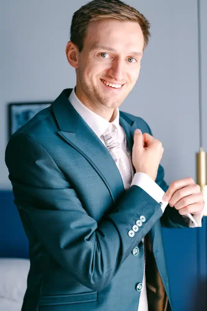 Lächelnder Bräutigam im blauen Anzug mit heller Krawatte richtet seinen Hemdsärmel, Getting Ready zur Hochzeit in der Orangerie Eisenstadt