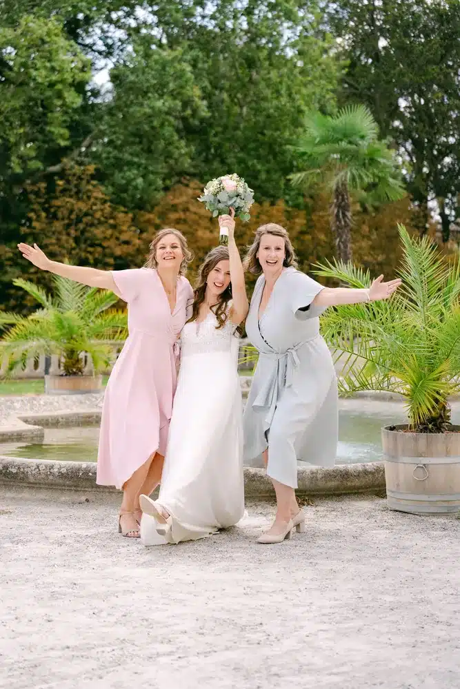 Braut jubelt mit zwei Frauen in Rosa und Hellblau vor den Palmen im Garten der Orangerie im Schlosspark Eisenstadt