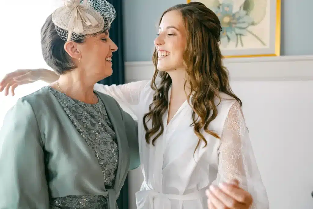 Braut im weißen Morgenmantel lacht mit einer Begleiterin im grünen Kleid und Fascinator beim Getting Ready zur Hochzeit im Schlosspark Eisenstadt