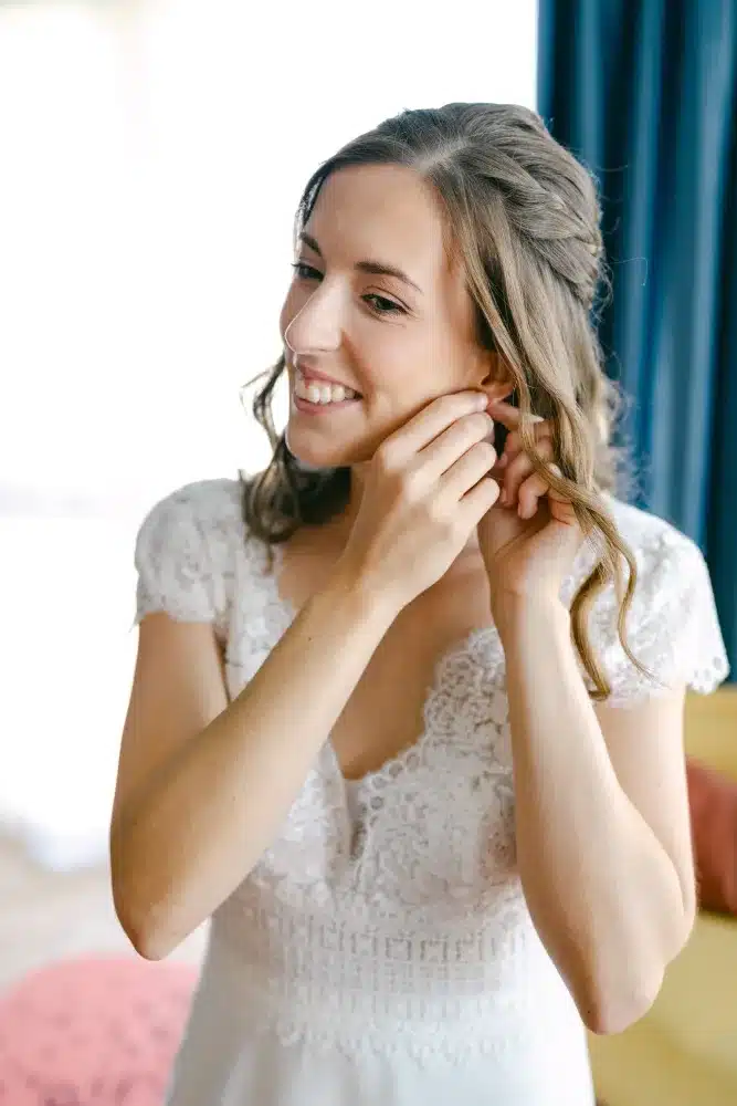Lächelnde Braut im Spitzenkleid legt ihren Ohrring an, Getting Ready vor der Hochzeit in der Orangerie im Schlosspark Eisenstadt
