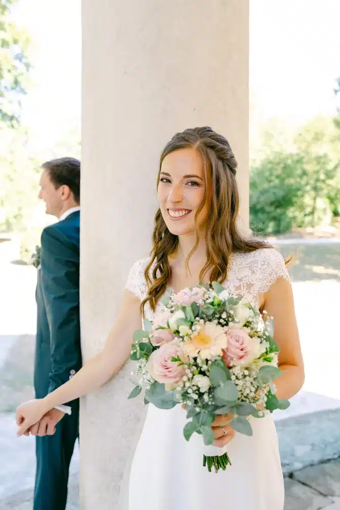 Lächelnde Braut mit Brautstrauß aus Rosen und Eukalyptus vor einer Säule, der Bräutigam im Hintergrund, Hochzeit im Schlosspark Eisenstadt