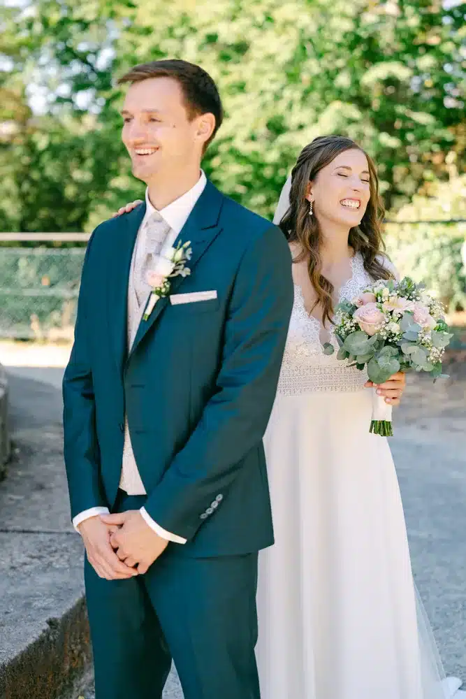 Braut mit Brautstrauß steht lachend hinter dem Bräutigam, der noch nach vorne blickt, First Look vor der Hochzeit im Schlosspark Eisenstadt