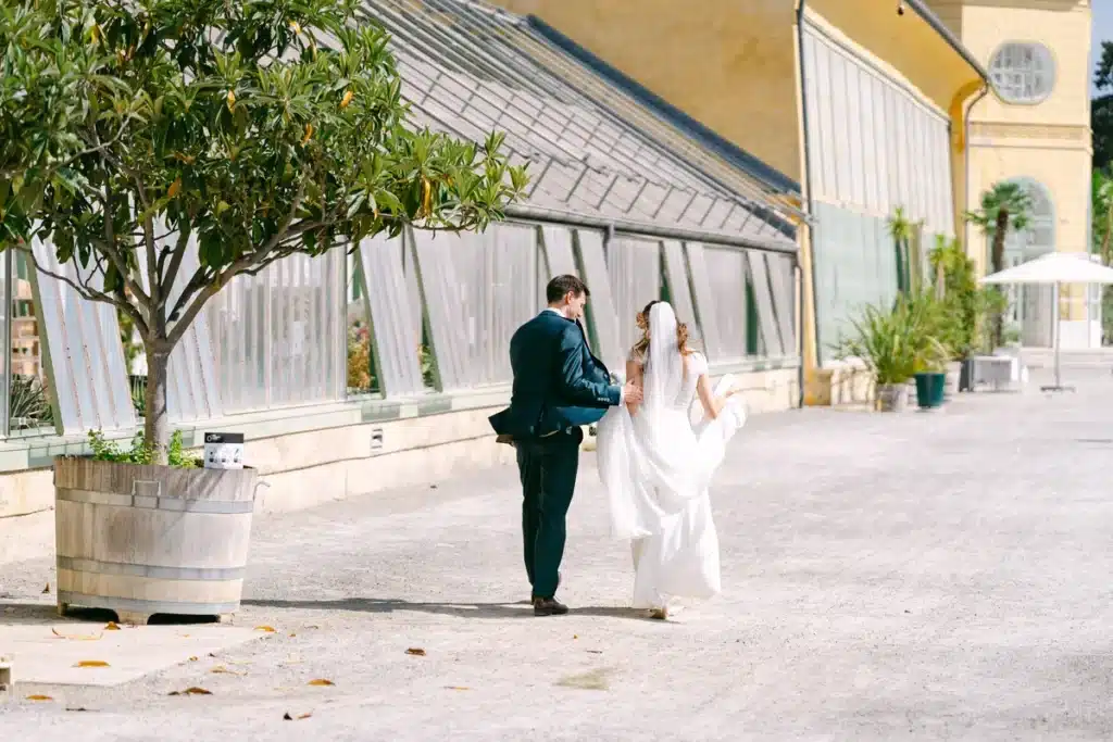 Brautpaar geht Hand in Hand entlang der Glasfassade der Orangerie im Schlosspark Eisenstadt, im Vordergrund Pflanzenkübel