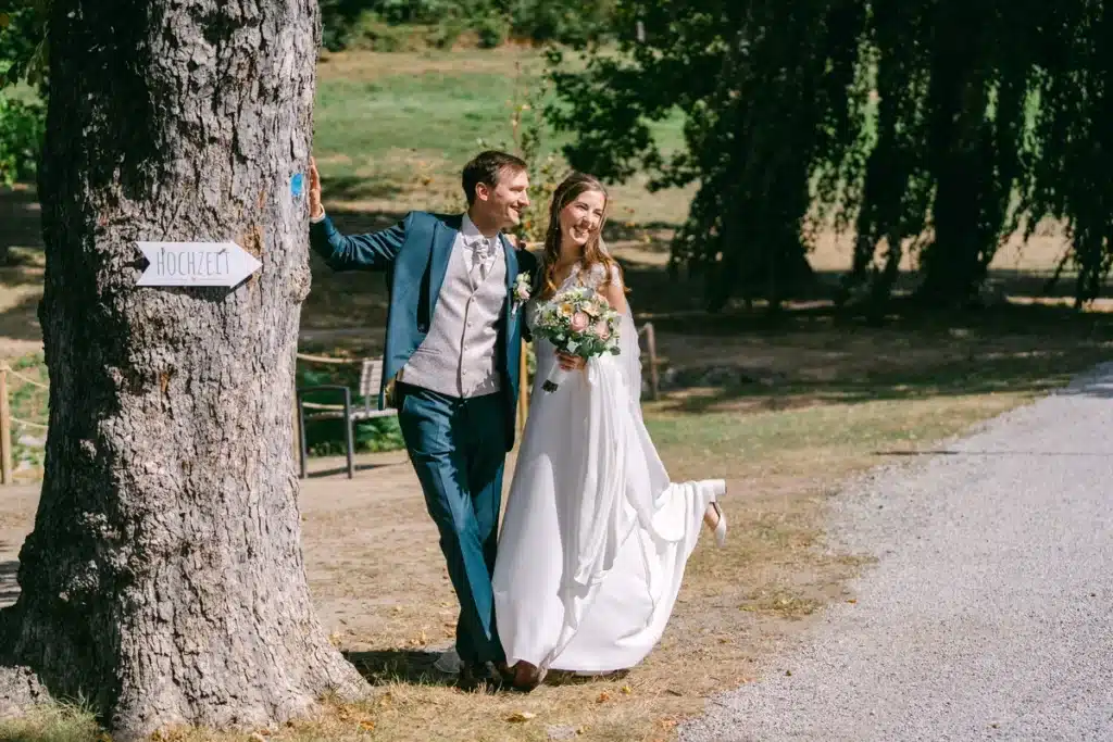 Brautpaar geht neben einem mächtigen alten Baumstamm durch den Schlosspark Eisenstadt, Paarfotos am Hochzeitstag