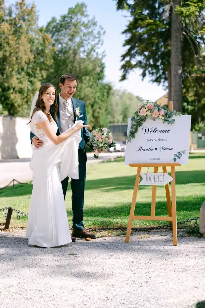 Brautpaar steht neben dem Welcome-Schild auf einer Holzstaffelei im Park, Hochzeit in der Orangerie im Schlosspark Eisenstadt