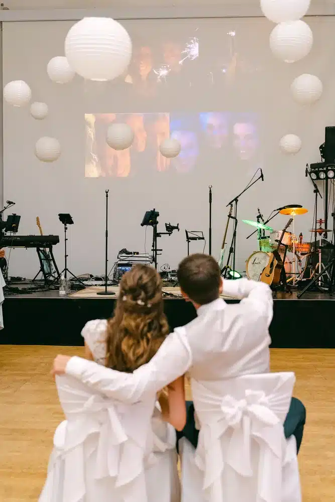 Brautpaar sitzt eng beieinander und blickt auf eine Leinwand vor der Bühne im Festsaal der Orangerie Eisenstadt, darüber weiße Papierlampions