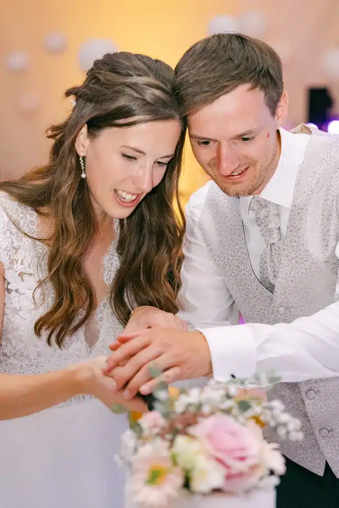 Brautpaar schneidet gemeinsam die Hochzeitstorte an, beide blicken lachend nach unten, Hochzeitsfeier in der Orangerie Eisenstadt