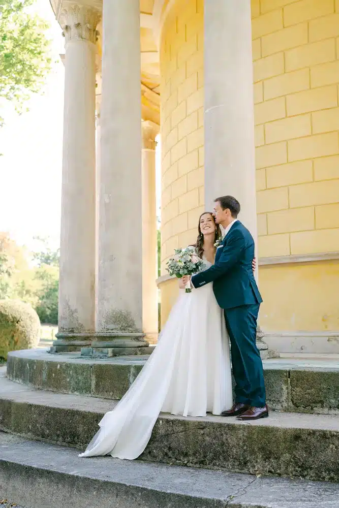Brautpaar umarmt sich auf den Steinstufen des gelben Säulentempels, Paarfotos beim Heiraten in der Orangerie im Schlosspark Eisenstadt