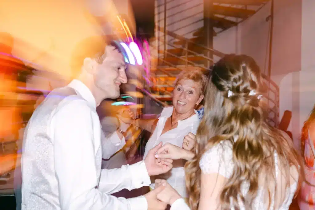 Brautpaar und ein Gast lachen gemeinsam auf der Tanzfläche im bunten Licht, Hochzeitsparty in der Orangerie Eisenstadt