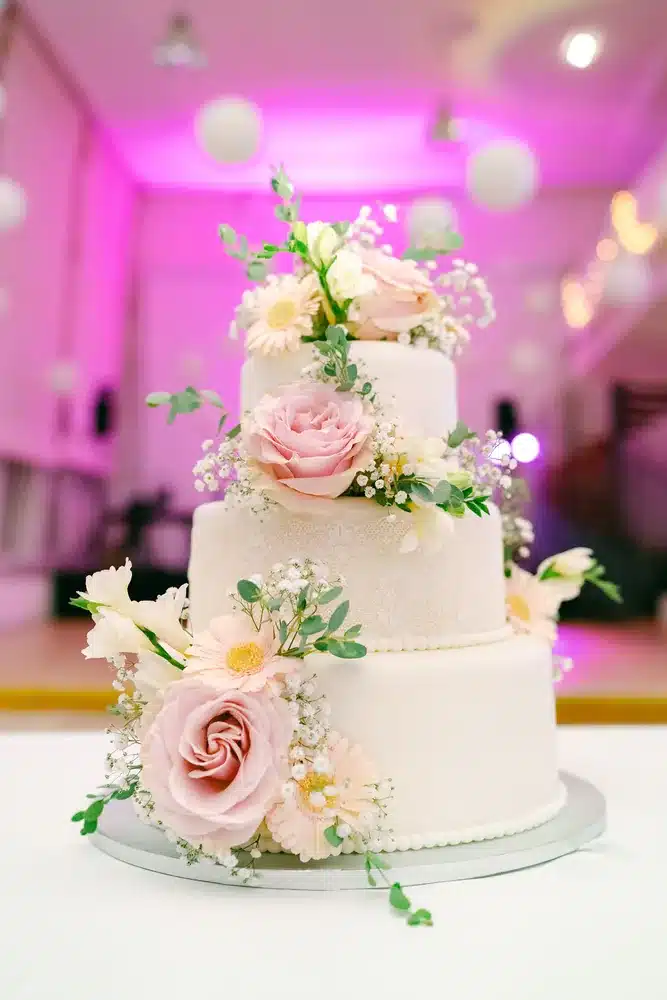 Dreistöckige weiße Hochzeitstorte mit rosafarbenen Rosen und Blüten im violetten Licht des Festsaals der Orangerie Eisenstadt