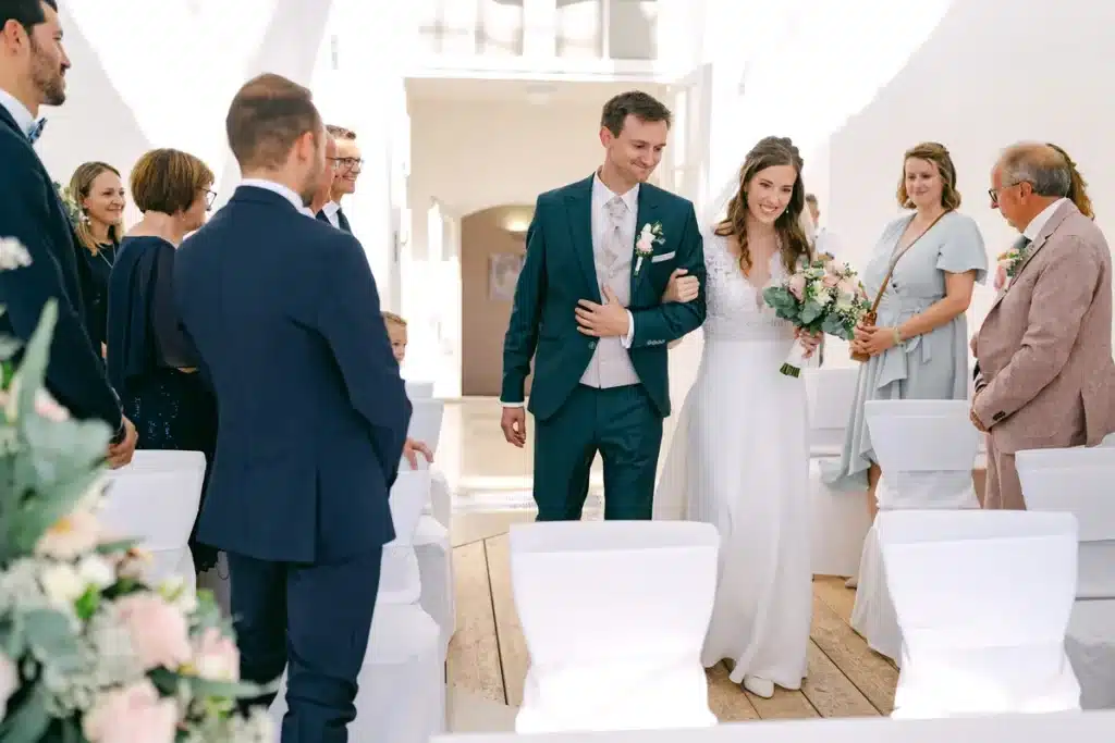 Brautpaar zieht Hand in Hand zwischen den stehenden Gästen in den Trausaal der Orangerie ein, Heiraten in der Orangerie im Schlosspark Eisenstadt