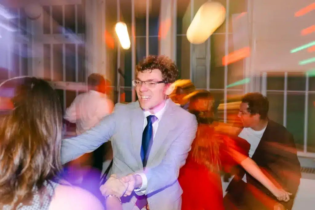 Hochzeitsgast mit Brille und hellem Sakko tanzt ausgelassen zwischen anderen Gästen im bunten Partylicht der Orangerie Eisenstadt