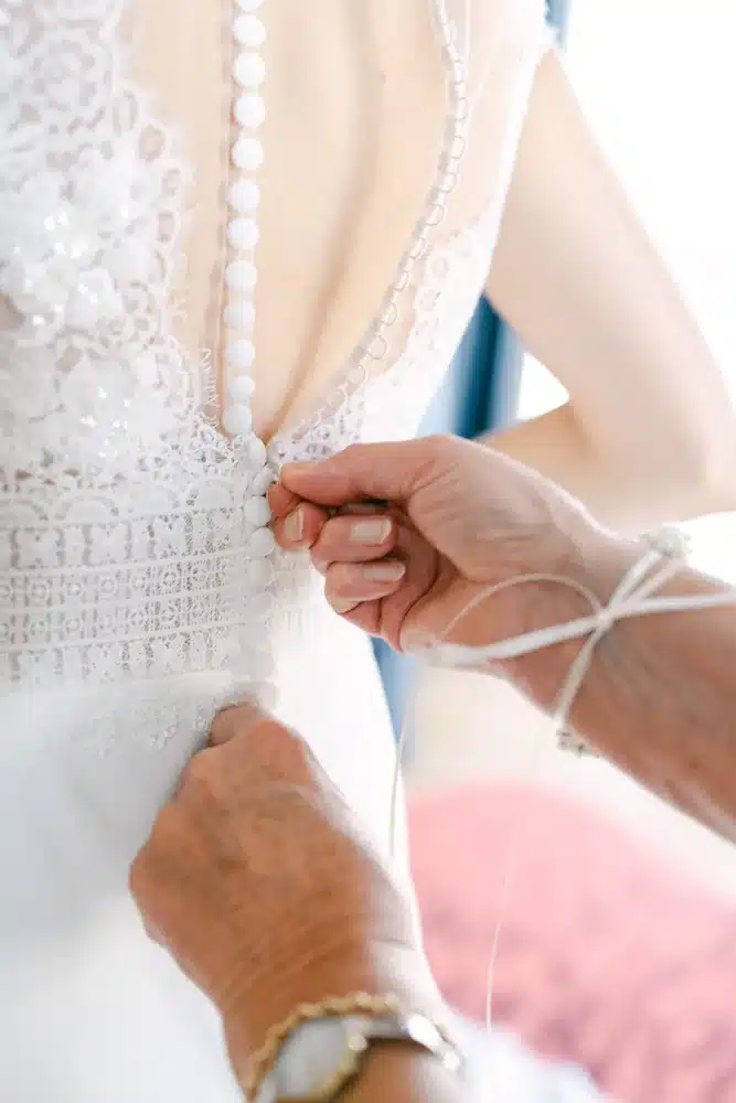Hände schließen die Knopfreihe am Rücken des Spitzenbrautkleids, Getting Ready zur Hochzeit in der Orangerie im Schlosspark Eisenstadt