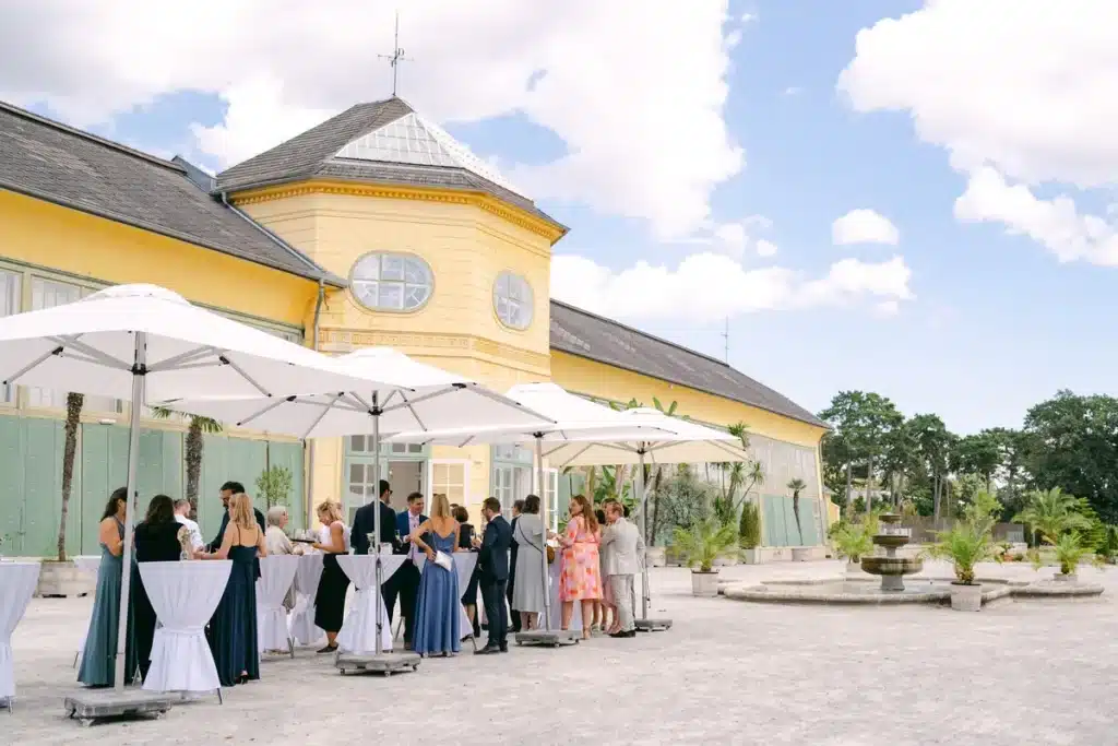 Hochzeitsgäste beim Sektempfang unter weißen Sonnenschirmen auf dem Kiesplatz vor der gelben Orangerie im Schlosspark Eisenstadt