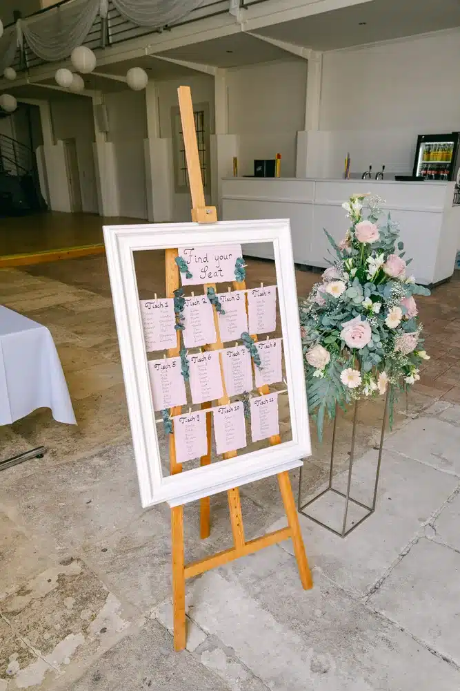 Sitzplan der Hochzeit auf einer Holzstaffelei neben einem Blumengesteck mit Rosen vor dem Festsaal der Orangerie Eisenstadt