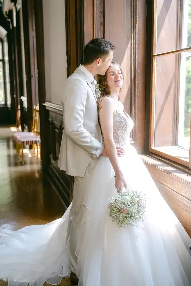 Heiraten in der Hochzeitslocation Schloss Margarethen am Moos – Getting Ready: Bräutigam küsst die Braut auf die Stirn vor dem Fenster