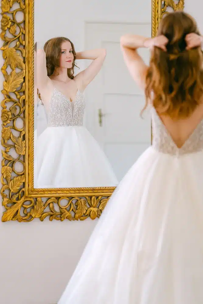 Heiraten in der Hochzeitslocation Schloss Margarethen am Moos – Getting Ready: Braut bindet sich vor einem goldgerahmten Spiegel die Haare hoch