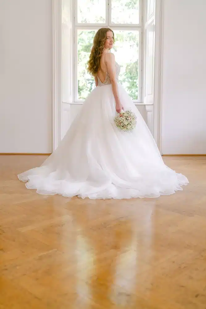 Heiraten in der Hochzeitslocation Schloss Margarethen am Moos – Brautpaarshooting: Braut steht mit Strauß am Fenster und blickt über die Schulter
