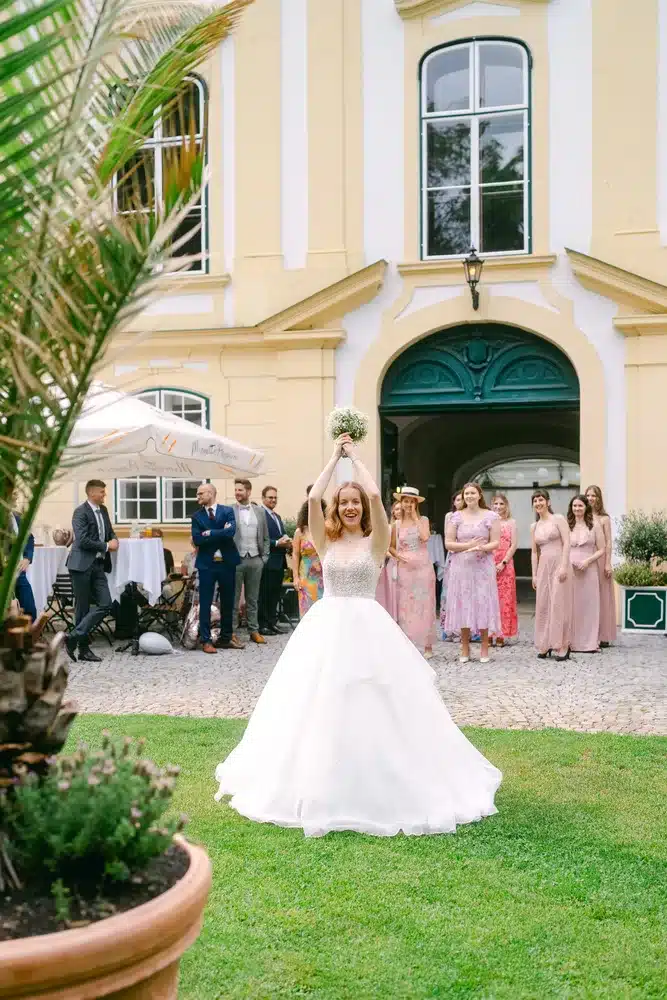 Heiraten in der Hochzeitslocation Schloss Margarethen am Moos – Brautpaarshooting: Braut wirft den Brautstrauß vor der Schlossfassade