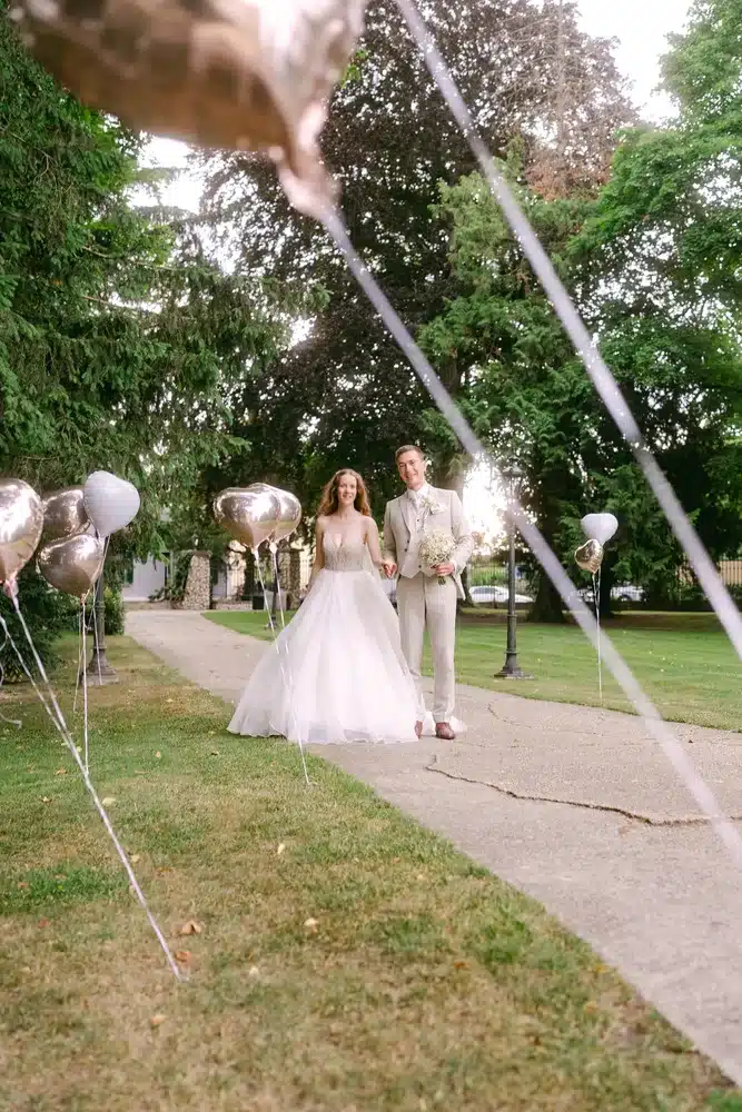 Heiraten in der Hochzeitslocation Schloss Margarethen am Moos – Brautpaarshooting: Brautpaar läuft durch einen Weg mit Herzluftballons