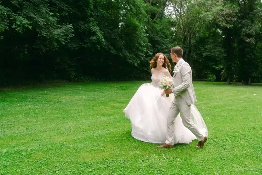 Heiraten in der Hochzeitslocation Schloss Margarethen am Moos – Brautpaarshooting: Brautpaar läuft lachend über eine Wiese