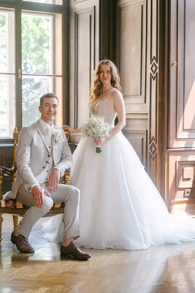 Heiraten in der Hochzeitslocation Schloss Margarethen am Moos – Brautpaarshooting: Brautpaar posiert auf einem Barockstuhl im Holzsaal