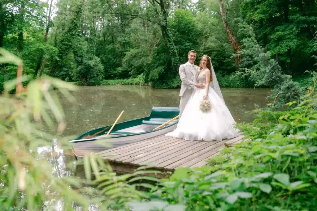 Heiraten in der Hochzeitslocation Schloss Margarethen am Moos – Brautpaarshooting: Brautpaar steht am See neben einem Ruderboot
