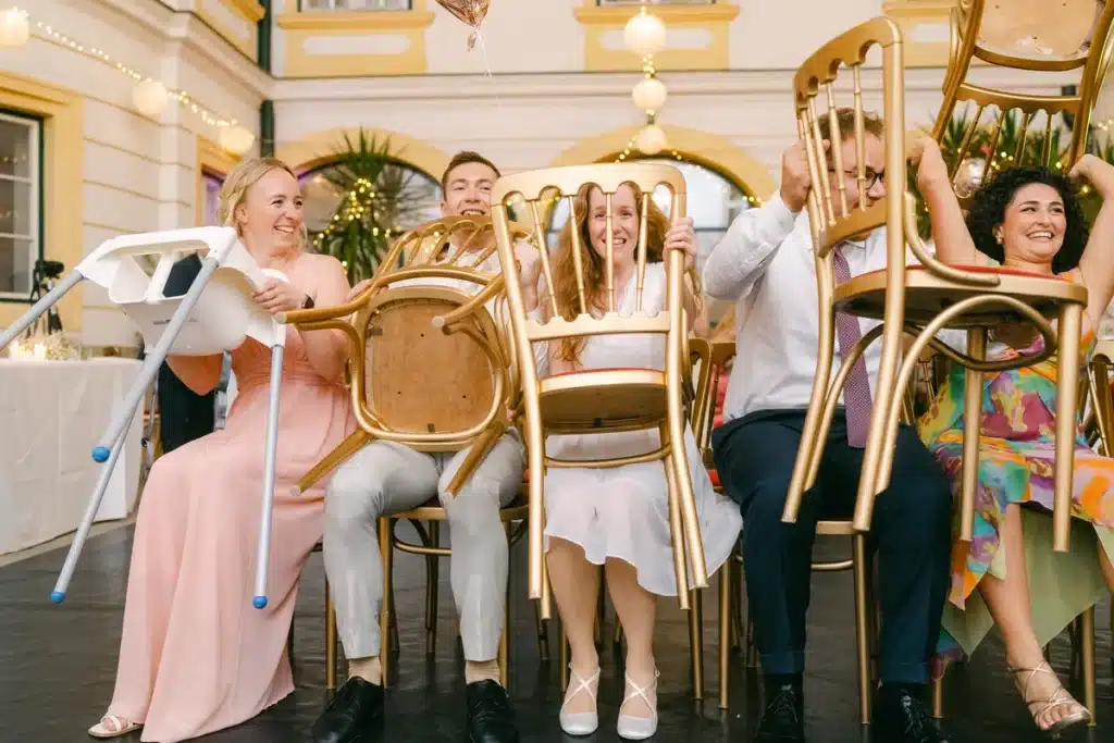Heiraten in der Hochzeitslocation Schloss Margarethen am Moos – Feier, Hochzeitstanz und Party: Gäste heben lachend goldene Stühle hoch