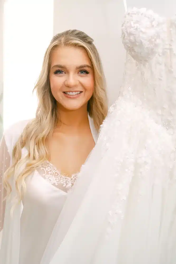 Lächelnde Braut im weißen Morgenmantel steht neben ihrem hängenden Brautkleid