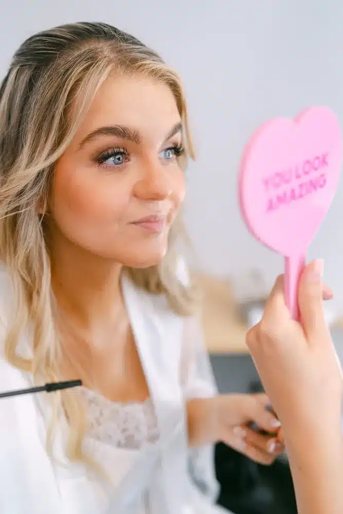 Braut beim Make-up hält einen rosa Herzspiegel mit der Aufschrift You Look Amazing