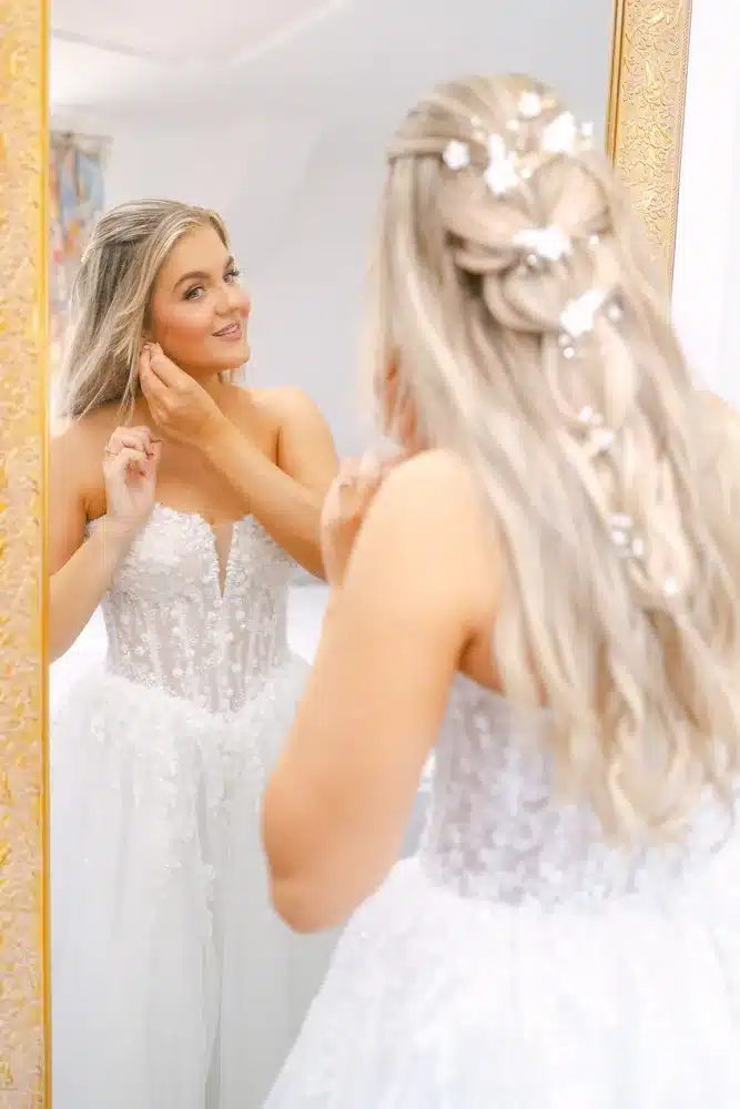 Braut im Spitzenkleid mit Haarschmuck betrachtet sich im goldgerahmten Spiegel