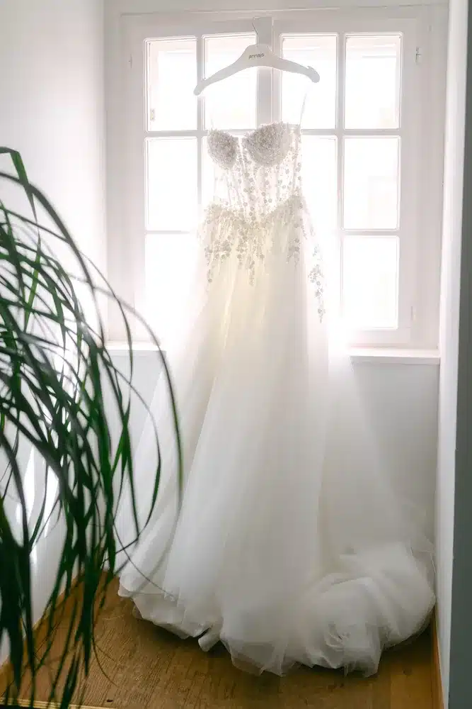 Weißes Brautkleid mit Spitzencorsage hängt am Fenster im hellen Gegenlicht