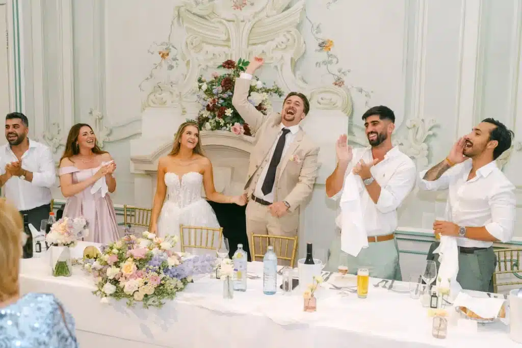 Brautpaar und Gäste jubeln mit erhobenen Armen am Brauttisch im Festsaal