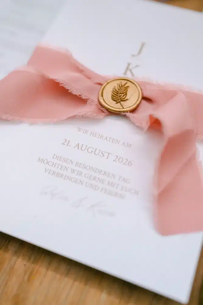 Hochzeitseinladung mit rosa Seidenband und goldenem Wachssiegel auf einem Holztisch