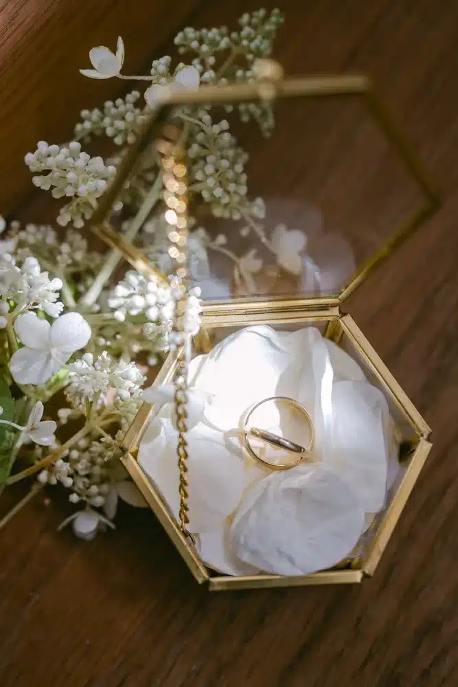 Zwei Eheringe in einer sechseckigen Ringbox aus Glas mit weißen Blüten
